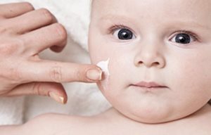 La mano de un adulto aplicándole crema al rostro de un bebé.